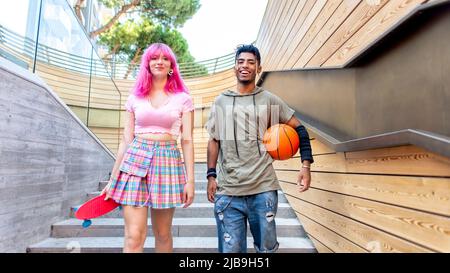 Alternative verschiedene Paar hängen zusammen chillend und lächelnd zu Fuß in der Stadt. Glückliche interracial Freunde halten Skateboard und Basketball Stockfoto