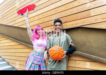 Alternative diverse paar hängen zusammen chillend und lächelnd stehen vor der Hintergrundwand. Glückliche interracial Freunde halten Skateboard an Stockfoto