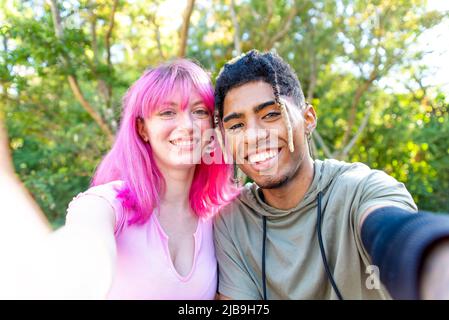 Alternative diverse paar hängen in einem Park machen ein pov Selfie-Porträt halten Smartphone auf beiden Seiten. Interracial Teenager Freunde entspannen Stockfoto