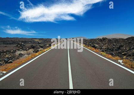 Lanzarote - Timanfaya NP: Fahrt auf endlos leerer Asphaltstraße zwischen schwarzen Lavasteinen in karger, rauer Landschaft bis zum Horizont, blauem Himmel, weiß Stockfoto