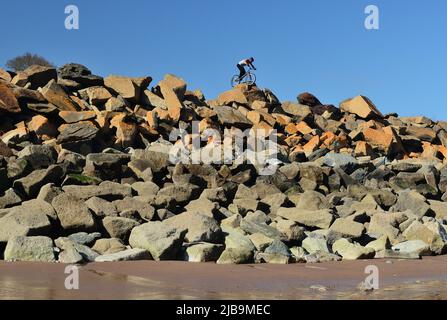 Ein akrobatischer Radfahrer, der Stunts auf mehrfarbigen Felsenpanzern auf der Meeresabwehr ausführt, vom Strand unten bei Ebbe gesehen. Stockfoto