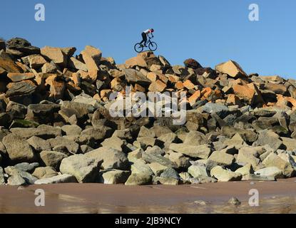 Ein akrobatischer Radfahrer, der Stunts auf mehrfarbigen Felsenpanzern auf der Meeresabwehr ausführt, vom Strand unten bei Ebbe gesehen. Stockfoto