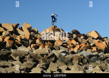 Ein akrobatischer Radfahrer, der Stunts auf mehrfarbigen Felsenpanzern auf der Meeresabwehr ausführt, vom Strand unten bei Ebbe gesehen. Stockfoto