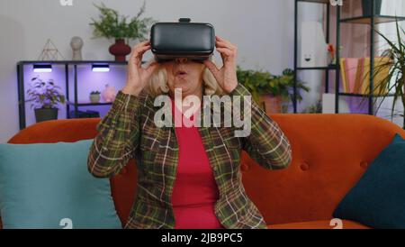 Ältere ältere Frau mit Headset Helm App, um Simulationsspiel zu spielen, Simulation Brille Gadget-Technologie. Virtual Reality 3D 360-Video ansehen. Reife Großmutter in VR-Brille auf der Couch zu Hause Stockfoto