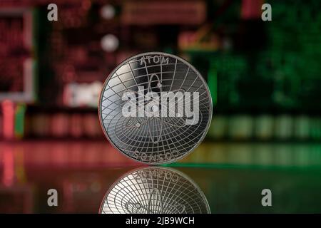 Cosmos ATOM Cryptocurrency Physical Coin platziert auf reflektierender Oberfläche mit Mikroschema im Hintergrund und beleuchtet mit grünen und roten Lichtern. Stockfoto