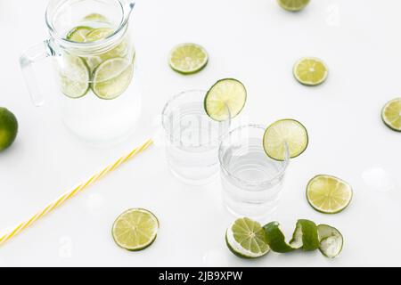 Auf einem weißen Tisch befindet sich ein Glaskrug Limonade, daneben zwei Gläser gefüllt mit Limonade und vielen Limonen. Stockfoto