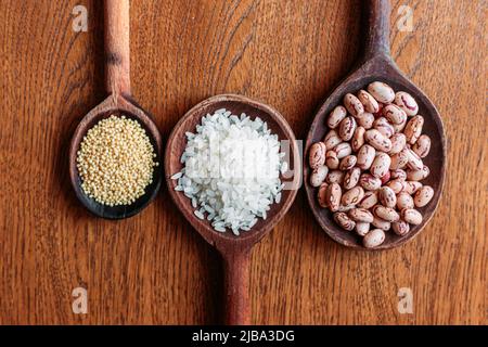 Weißer Reis, Pinto-Bohnen und Proso-Hirse-Samen auf hölzernen Küchenlöffeln. Brauner, hölzerner Hintergrund. Glutenfreie, gesunde Lebensmittel Stockfoto