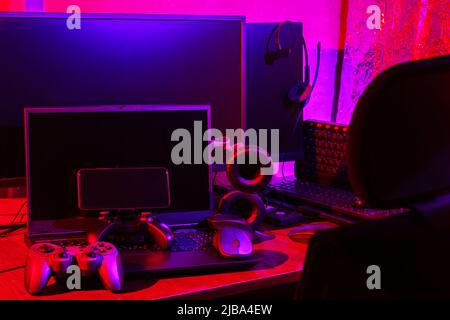 Arbeitsbereich des Spielers. Gamepads, Computermaus, Kopfhörer, Tastatur und Laptop auf dem Tisch. Gaming-Zubehör und Computer-Ausrüstung in Neonlicht. Ga Stockfoto