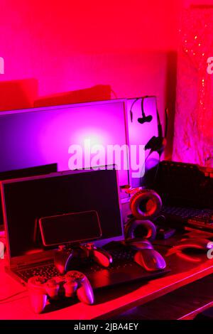 Arbeitsbereich des Spielers. Gamepads, Computermaus, Kopfhörer, Tastatur und Laptop auf dem Tisch. Gaming-Zubehör und Computer-Ausrüstung in Neonlicht. Ga Stockfoto