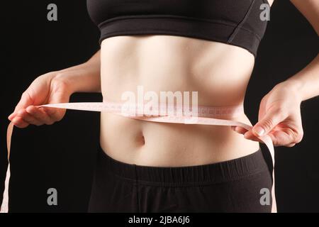 Eine Frau misst den Umfang ihres Bauches auf schwarzem Hintergrund. Das Konzept der Gewichtsabnahme Stockfoto