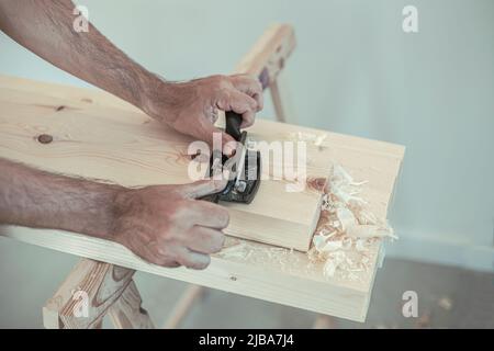 Zwei Hände eines Mannes, der in einer Werkstatt mit natürlichem Licht an einer Holzlatte mit einem Schrankkratzer arbeitet Stockfoto