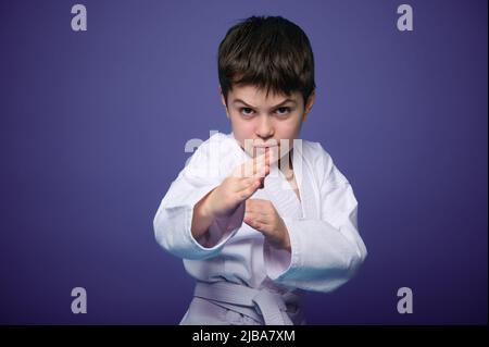 Taillenlänge Porträt eines selbstbewussten starken dunkelhaarigen kaukasischen Kindes, Aikido Kämpfer üben Kampfkünste auf lila Hintergrund mit Werbung Stockfoto