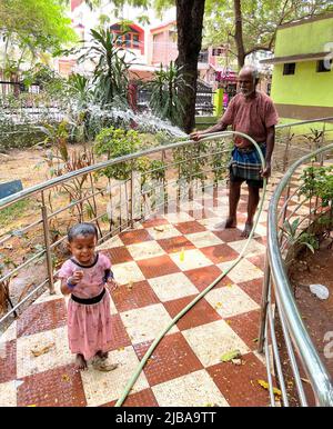 Chennai, Indien, 4.. Juni 2022: Ein Gärtner (Großvater), der die Pflanzen in einem Park wässert, während seine Enkelin den Nieselregen genießt, der die Hitze in Chennai schlägt. Seshadri Sukumar Stockfoto