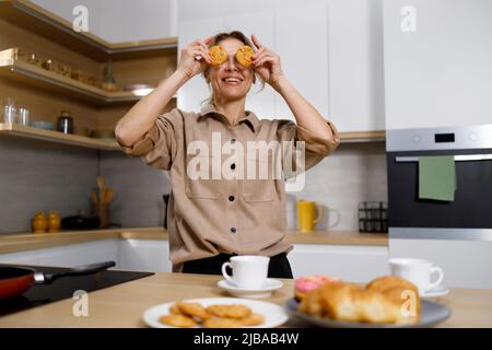 Schöne blonde Frau hält Cookies auf den Augen. Weibliche Spaß in der Küche beim Tee Stockfoto