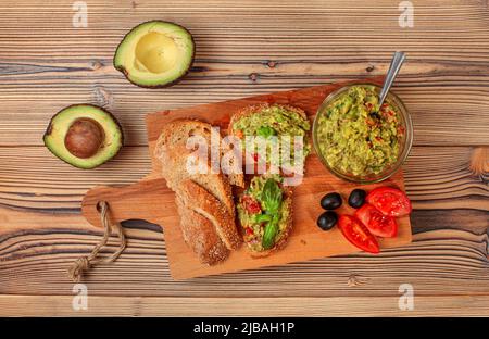 Frisch zubereitete Guacamole in kleiner Glasschüssel, Brot, Tomaten, Oliven am Arbeitsbrett und zwei Avocados-Hälften weiter, Blick von oben Stockfoto