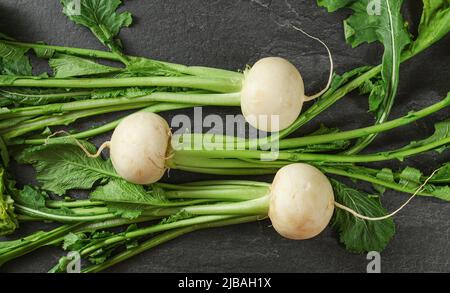 Drei weiße Rettich-Birnen mit grünen Blättern auf schwarzem schieferartigen Brett, Blick von oben Stockfoto