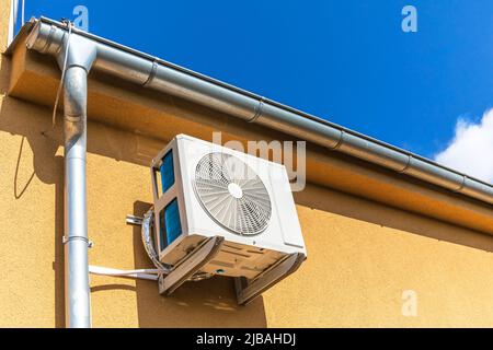 Heißer Sommertag. Kühlung. Klimaanlage, die im Freien an der Wand eines modernen Gebäudes oder Hauses neben dem Dach gegen einen blauen Himmel aufgehängt ist. Stockfoto