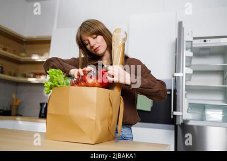 Die junge Frau kam mit einem vollen Paket von Produkten vom Einkaufen zurück und stellte sie in einen leeren Kühlschrank. Hausfrau nimmt Gemüse aus einem pa Stockfoto