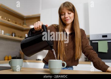 Junge Frau gießt heißes Wasser aus dem Wasserkocher in den Becher in der Küche. Mädchen macht Tee Stockfoto