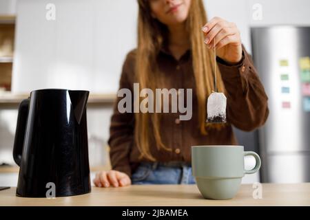 Junge Frau macht Tee in der Küche. Mädchen steckt einen Teebeutel in ein Glas. Schwarzer Wasserkocher auf dem Tisch. Stockfoto