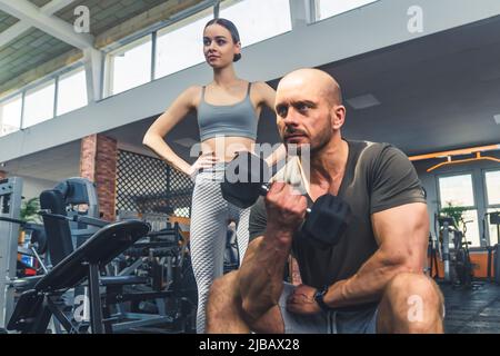 Kaukasisches Fitnesspaar im modernen Fitnessstudio. Muskulös starke Glatze Heben Hantel mit einer Hand, während auf einer Bank sitzen. Junges Mädchen in Sportkleidung, das ihn im Spiegel ansieht. Hochwertige Fotos Stockfoto