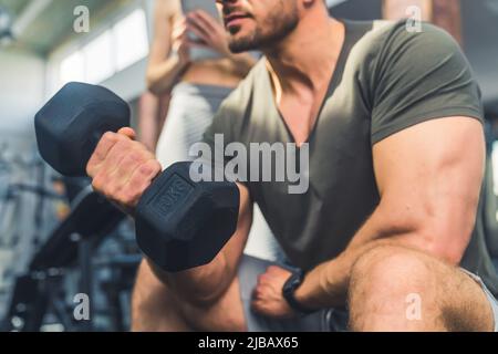 Closeup Shop im Gym Interieur eines starken Bizeps. Kaukasischer Bodybuilder Kerl und seine große schwarze Hantel. Hochwertige Fotos Stockfoto