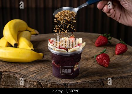 Köstliche brasilianische Açaí-Creme, in einem Plastikbecher mit Erdbeere, Banane und Granola Topping, auf einem rustikalen Holzhintergrund. Sommer acai Smoothie Stockfoto