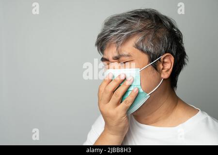 Kranker asiatischer Mann, der eine medizinische Gesichtsmaske trägt und hustet und seinen Mund mit meiner Hand bedeckt. Konzept zum Schutz des pandemischen Coronavirus und Atemschutzgeräts Stockfoto
