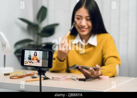 Junge asiatische Frau vloggt sich über Beauty-Produkte mit einem Video für ihren Blog über Kosmetik mit Handy-Kamera zu Hause machen. Stockfoto