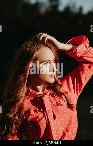 Portait der Schönheit junge rothaarige Frau mit langen lockigen dicken Haaren in rotem Hemdkleid mit weißen Punkten posiert auf einer Kamera im Sonnenlicht gekleidet Stockfoto