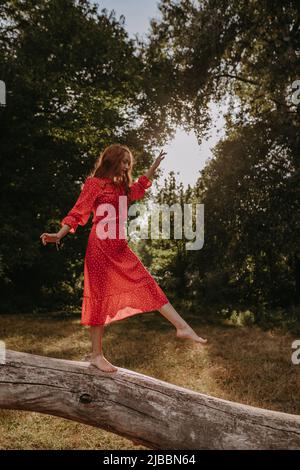 Rothaarige junge Frau im roten Sommerlandkleid balanciert und tanzt auf einem trocken gefallenen Baum mitten im Wald Stockfoto