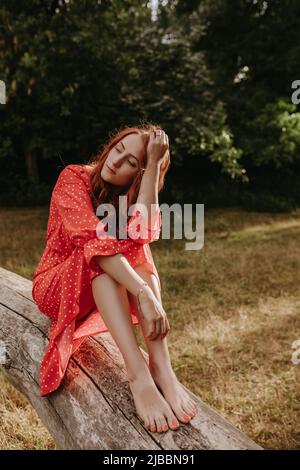 Hübsche junge Erwachsene Frau in einem roten Kleid mit weißen Punkten mit roten Haaren sitzt auf einem trocken gefallenen Baum und posiert auf einer Kamera. Weibliche Modell berühren sie h Stockfoto