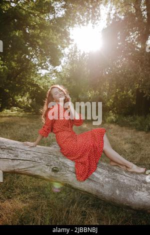 Hübsche junge Erwachsene Frau in einem roten Kleid mit weißen Punkten mit roten Haaren sitzt auf einem trocken gefallenen Baum und posiert auf einer Kamera. Weibliche Modell berühren Gesicht Stockfoto