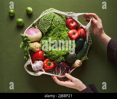 Frauenhände halten einen ökologischen Baumwollstring-Beutel mit frischem Obst und Gemüse auf grünem Hintergrund - Zero Waste Konzept Stockfoto