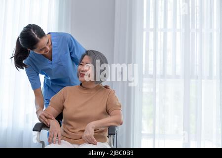 Junge Krankenschwester kümmert sich um ältere behinderte Frau, die im Rollstuhl mit Kopierraum sitzt. Eine weibliche Unterstützerin, die einer älteren Patientin hilft, während sie lächelt Stockfoto