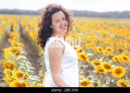 Weicher Fokus auf Porträt von lächelndem Mädchen im Sonnenblumenfeld. Frau mit lockigen Haaren in weißem Kleid geht. Sonnenblume ist ukrainische Landwirtschaft. Gelbgold Stockfoto