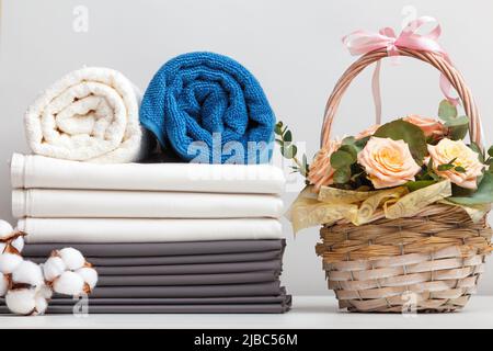 Ein Stapel Bettwäsche Bettwäsche Bettwäsche Brötchen mit Handtüchern. Korb mit Rosenblüten auf dem Tisch. Stockfoto