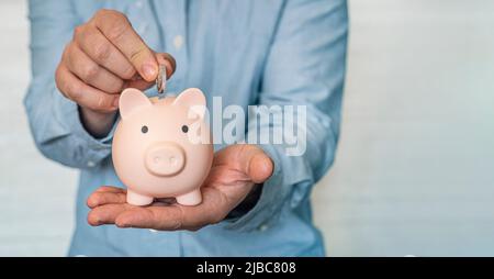 Geld in ein Sparschwein stecken. Ein junger Geschäftsmann in einem blauen Hemd steckt eine Münze in das Sparschwein. Das Konzept des Sparens. Platz kopieren Stockfoto