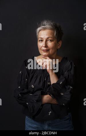 Wunderschöne reife Geschäftsfrau mit grauen Haaren posiert gelehntes Kinn auf ihre Hand isoliert auf schwarzem Hintergrund. Grau elegante Frau mit kurzen Haaren. Bild einer erfolgreichen reifen Frau. Gealterte Schönheit. Stockfoto