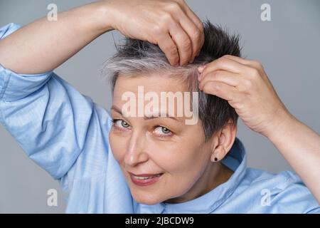 Hübsche Frau im Jahr 50s überprüft ihre Haare oder Schädelhaut im Spiegel. Schöne grauhaarige Frau, die mit Schuppen zu tun hat, Problem beim Einchecken im Spiegel. Gesundes Haar, Haarausfall Konzept. Stockfoto