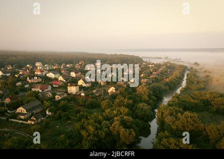 Private Wohnhäuser am Ufer des Flusses unter den Bäumen an einem nebligen Sommermorgen - Luftdrohne geschossen. Nebliger Morgen im Ökodorf: Häuser in der Nähe des Wassers zwischen dichten grünen Bäumen. Stockfoto