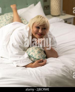 Luxus-Lifestyle-Shooting mit Plus-Size-Body-positive-Modell für Frauen, mit weißem Bademantel auf Kingsize-Bett Stockfoto