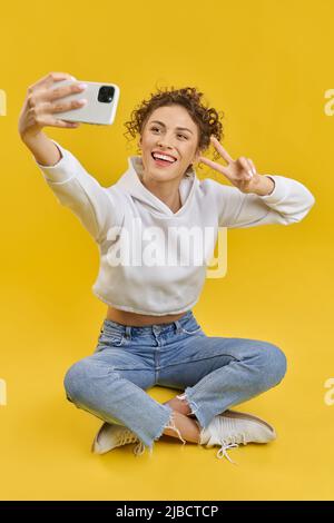 Hübsche Frau im Hoodie mit Siegesgeste und Selfie auf dem Boden mit dem Smartphone. Portrait-Ansicht von niedlichen Mädchen, das Foto, sitzt in Lotus-Pose, isoliert auf gelbem Hintergrund. Konzept von Selfie. Stockfoto