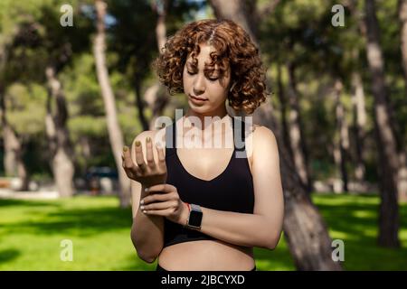 Die junge kaukasische Frau trägt einen schwarzen Sport-BH und steht auf dem Stadtpark und hält ihr schmerzhaftes Handgelenk im Freien. Leiden Schmerzen an Händen und Fingern, arthriti Stockfoto