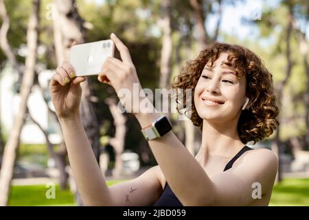 Junge Frau, die im Stadtpark mit ihrem Lächeln und ihrem Smartphone im Freien in sportlicher Kleidung lächelt und horizontale Fotos macht. Stockfoto