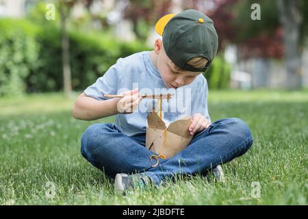 Kind, das auf dem Gras sitzt, auf der Straße Essen isst. Schmutzige Soße auf seinem T-Shirt. Nudeln fielen auf seine Kleidung. Draußen Stockfoto