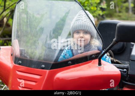 Vorderansicht eines dreijährigen kaukasischen Jungen, der auf dem Fahrersitz eines Quad-Bikes sitzt und durch die Windschutzscheibe blickt. Platz für Kopie auf der linken Seite. Stockfoto