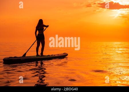 25. Juni 2021. Anapa, Russland. Sportliche Frau auf dem Paddelbrett am ruhigen Meer mit Sonnenuntergang oder Sonnenaufgang. Frau auf Red Paddle-Brett und warmen Sonnenuntergang Stockfoto