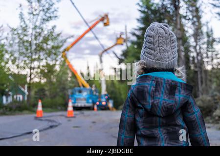 Rückansicht eines neugierigen, vierjährigen Jungen, der draußen steht, während die Powerline-Ingenieure im Hintergrund verschwommen sind, um den elektrischen Betrieb nach einem Sturm wiederherzustellen. Stockfoto