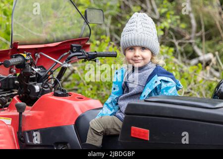 Porträt eines lächelnden vierjährigen Jungen im Freien, der einen grauen wolligen Hut und einen blauen Mantel trägt. Sitzen auf einem ländlichen Quad-Bike mit verschwommenem Waldhintergrund. Stockfoto